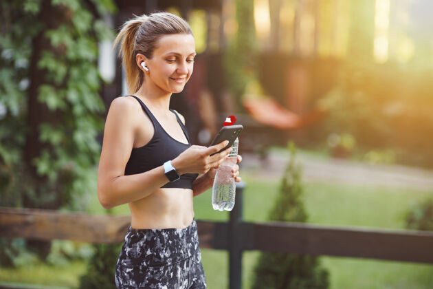 休息年轻女子在绿色公园的墙上锻炼后检查智能手机跑步瓶子水