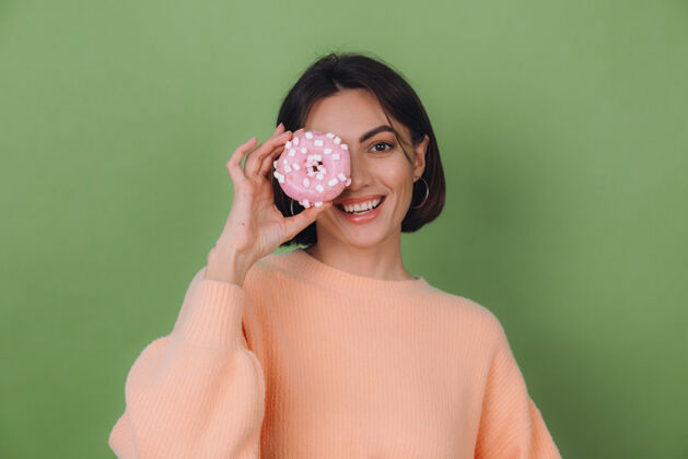 美味穿着休闲桃色毛衣的年轻时尚女性 隔离在绿色橄榄色墙壁上 粉色甜甜圈快乐复制空间女人年轻美味
