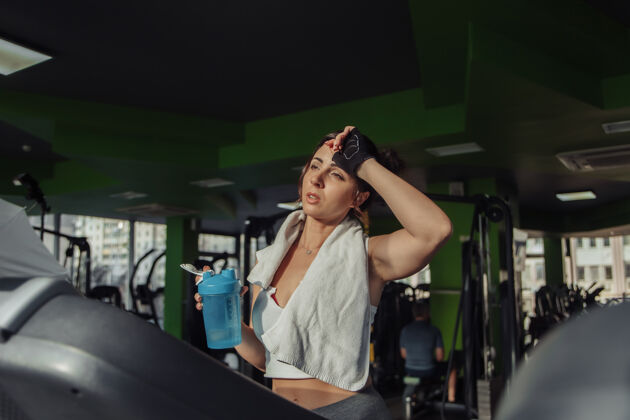 锻炼一个疲惫不堪的女人 肩上扛着毛巾 在跑步机上拿着水瓶减肥理念 有氧运动瓶子健身房俱乐部