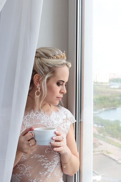 灯微笑的金发新娘在酒店房间喝咖啡光婚礼礼服