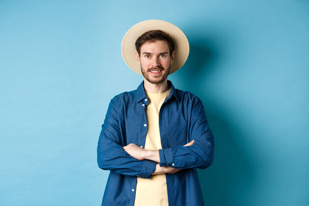 肖像一幅欢快的小伙子戴着草帽去度假的画像 双臂交叉在胸前 面带微笑 看上去很自信 站在蓝色的背景上人站胡子