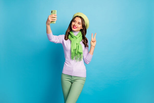 胜利搞笑迷人女士手持电话自拍照片显示v字标志穿时髦绿色贝雷帽紫色高领围巾裤子蓝色隔离墙拍摄社交乐趣