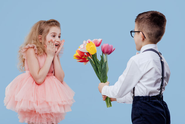 儿子两个快乐的孩子小男孩把花伸向一个孤立在蓝色墙上的小女孩期待奇迹花束
