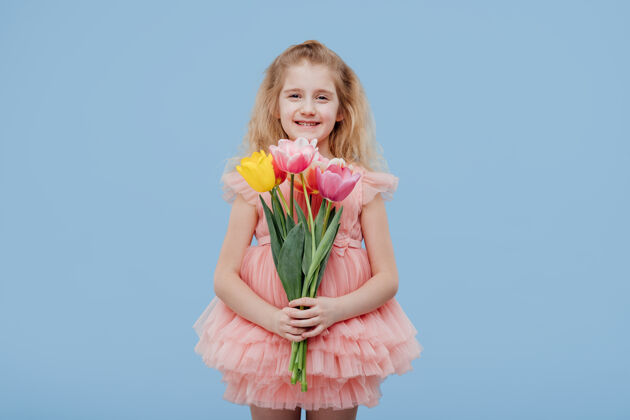 微笑快乐的小女孩穿着粉红色的裙子 手里拿着春天的花朵 孤零零地站在蓝色的墙上快乐孩子惊喜