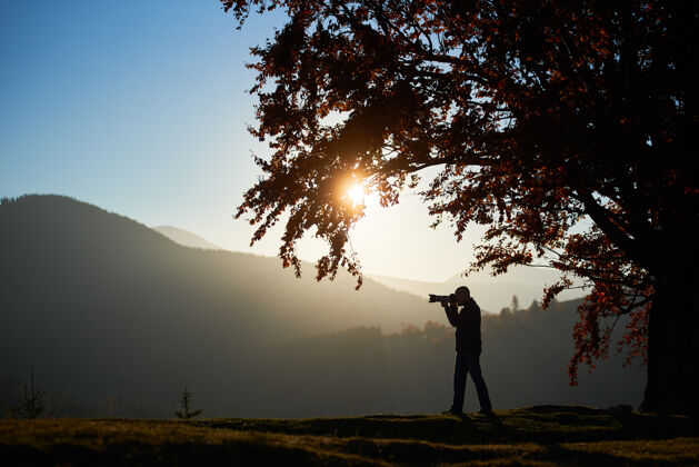 橡树徒步旅行者带着相机在大树下的山景背景下的草谷里森林人男性