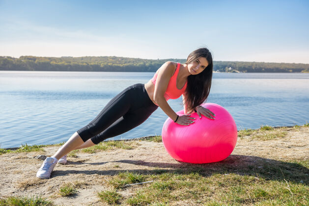 拉伸年轻柔韧的女子在湖边的粉红球上做早操运动健康运动员