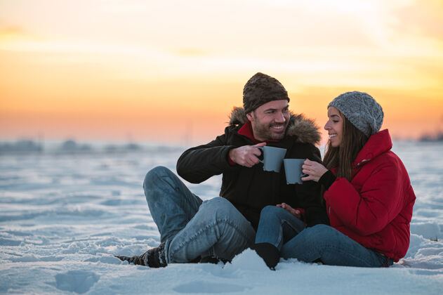 情侣积极的年轻人穿着暖和的外套 拿着热水瓶坐在雪地上 在乡下喝着热饮料内容关系远足