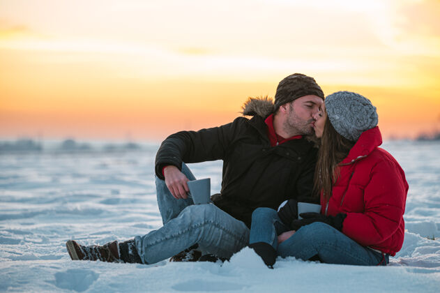 积极积极的年轻人穿着暖和的外套 拿着热水瓶坐在雪地上 在乡下喝着热饮料环境草地乡村