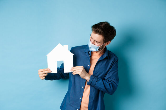 社会距离房地产和检疫概念戴着医学面具的年轻白种人站在蓝色背景上 看着纸屋的剪纸房子年轻控股