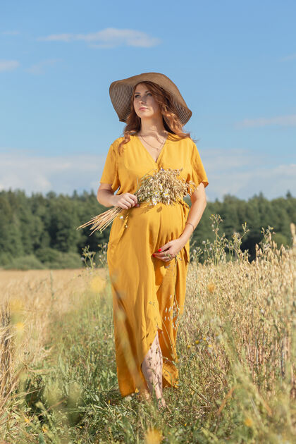 肚子在一个阳光明媚的夏日 一位年轻漂亮的孕妇穿着黄色的裙子 戴着黄色的帽子 走过一片麦田农村麦田阳光