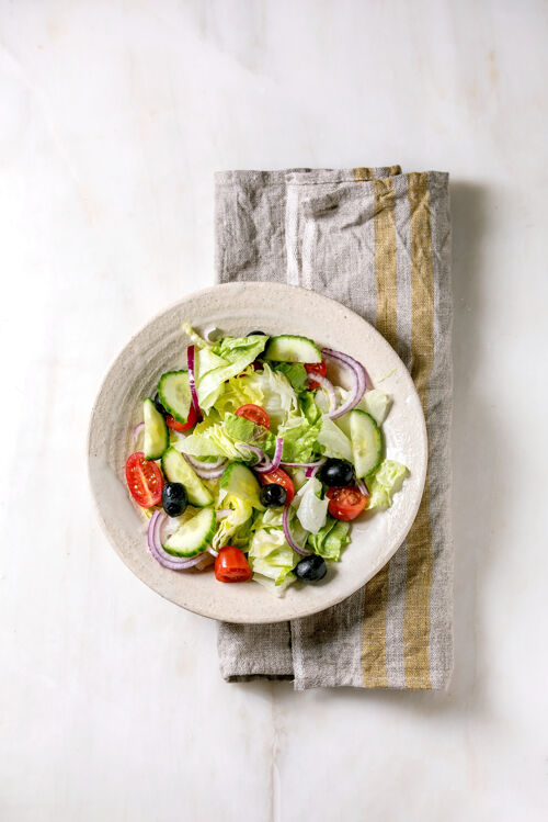 可口经典的蔬菜沙拉 配西红柿 黄瓜 洋葱 沙拉叶和黑橄榄 用白色陶瓷盘放在布餐巾上白色大理石背景平铺 复制空间洋葱膳食吃