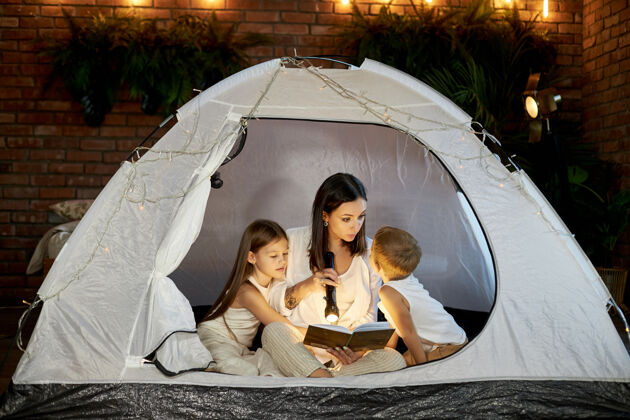 年轻妈妈坐在家里的帐篷里给孩子们读睡前故事母子俩抱在一起 手里拿着手电筒看书灯乐趣卧室