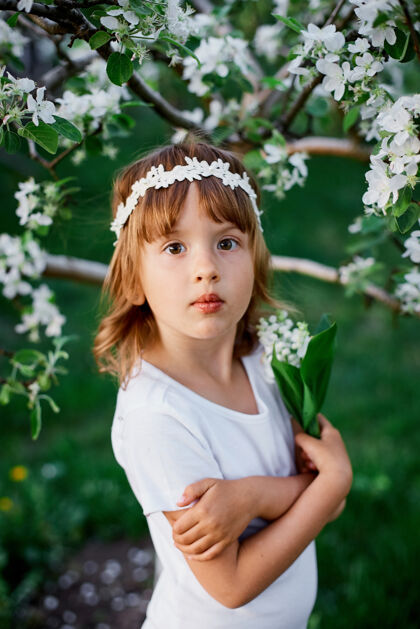 夏天可爱的5-6岁小女孩手持鲜花站在百花齐放的春天花园里 穿着白色的连衣裙 戴着花环在户外 春天来了乐趣童年夏装