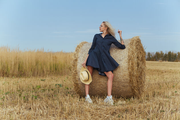田野一位年轻漂亮的女村民在一捆干草旁摆出一身衣服美丽草包帽子