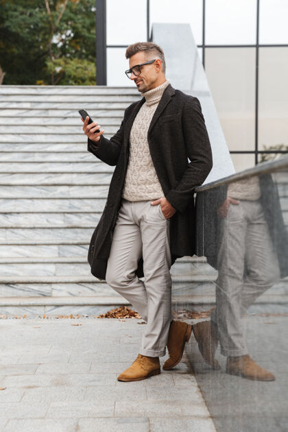 成熟30多岁的时尚男人戴着眼镜 穿过城市街道 使用智能手机的画像中心站立时尚