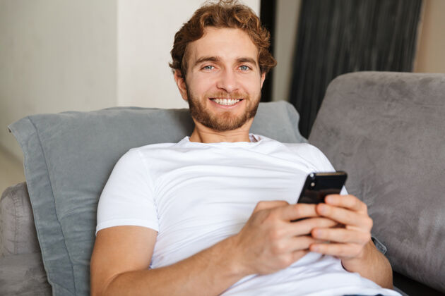 休闲一个英俊的留着胡子的年轻人在家里的沙发上用手机应用程序学习手机