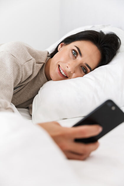 成熟30多岁的单身女性在家里穿着白色亚麻布躺在床上使用智能手机的照片放松公寓互动