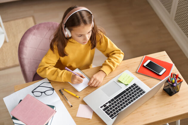 在线学习顶视图：戴着耳机的年轻女孩在她房间的办公桌上做作业学习观看办公桌