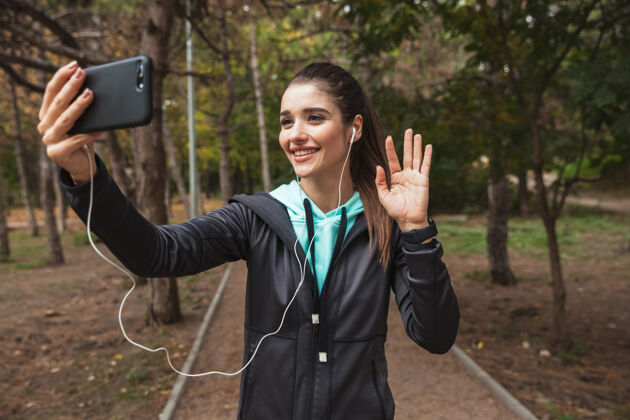 女士微笑的健身女士戴着耳机听音乐 站在公园里自拍运动耳机散步