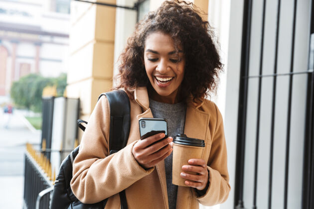 年轻快乐的非洲年轻女子穿着外套走在户外 拿着外卖咖啡杯 用手机风格外套手机