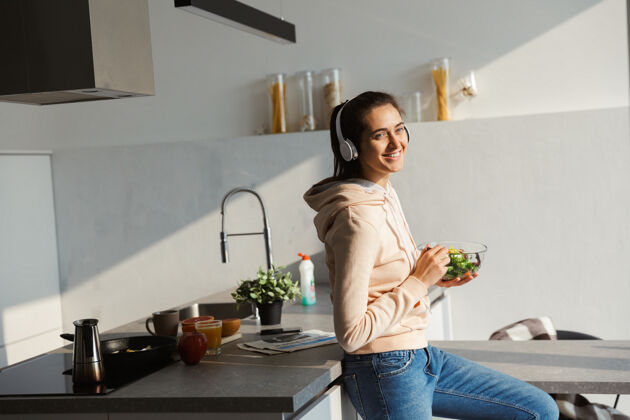 饮食快乐健康的女人每天早上站在厨房吃沙拉 戴着耳机听音乐休闲年轻早餐
