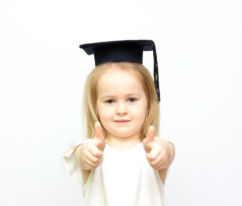 聪明在孤立的白色背景下 戴着学生帽的快乐小女生竖起大拇指好白人孩子