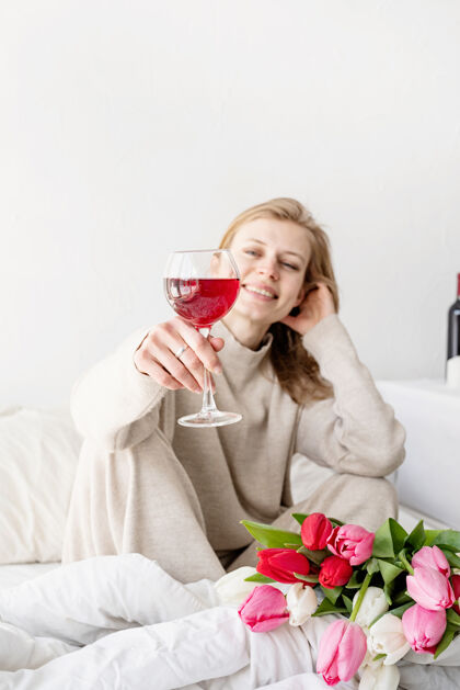 葡萄酒快乐的女人穿着睡衣坐在床上 愉快地欣赏着鲜花和一杯红酒浪漫卧室情绪