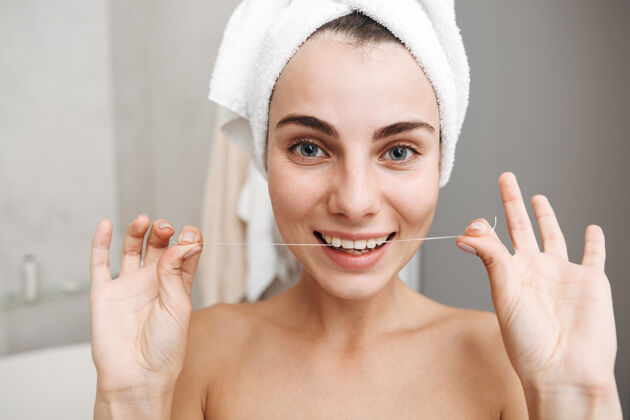 牙科特写镜头：一个头上戴着毛巾的年轻漂亮女人站在浴室里 用牙线日常年轻人清洁