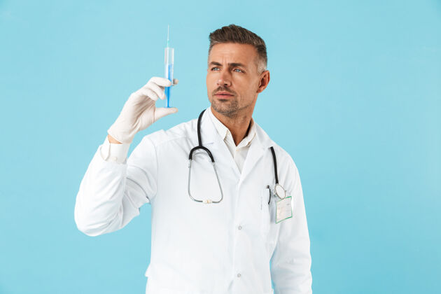 制服戴着听诊器戴着手套拿着注射器的白人医生的画像 孤立地站在蓝色的墙上医疗专家阳性