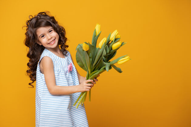 童年穿着条纹连衣裙的可爱小女孩手持一束郁金香在黄色的工作室里背景.spring妇女节活动漂亮花束