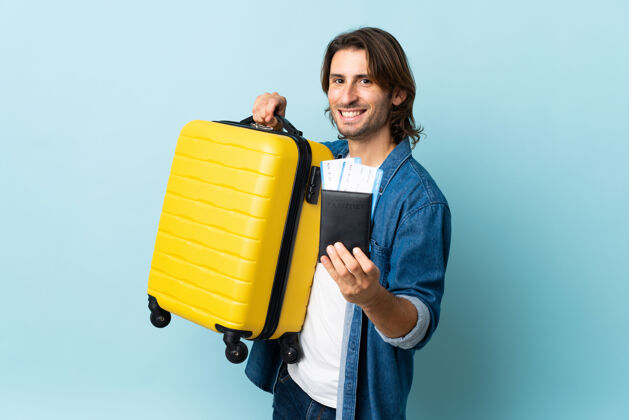 手提箱年轻帅哥带着手提箱和护照被隔离在蓝色的墙上度假包好长发