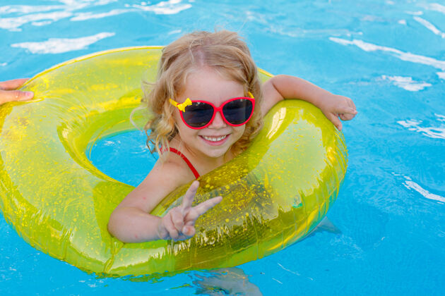 大笑夏天 一个戴着太阳镜和泳衣的小女孩在一个充气圈的游泳池里游泳眼镜飞溅阳光