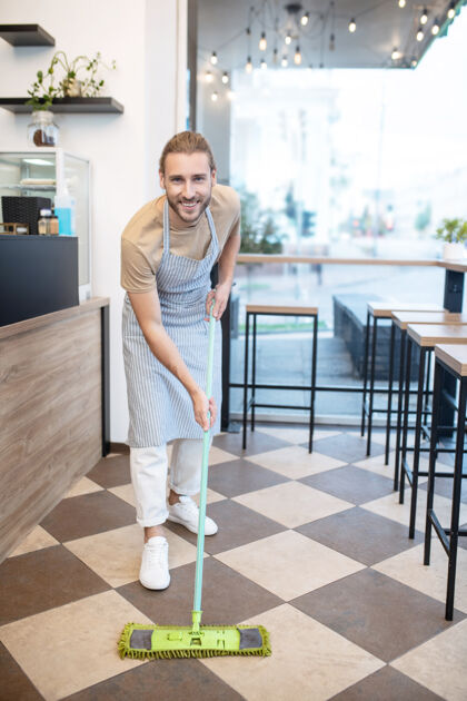 订单小商务微笑年轻的成年男子在围裙清洁地板在他的咖啡馆在白天家具全长整洁