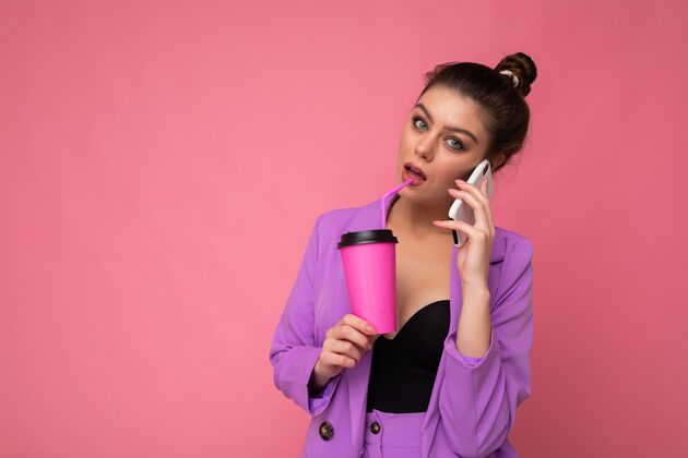 咖啡漂亮的黑发女人的肖像穿着时髦的紫色夹克 在智能手机上交谈 拿着纸杯里的外卖咖啡 隔离在粉色背景空白处做广告茶青年通讯