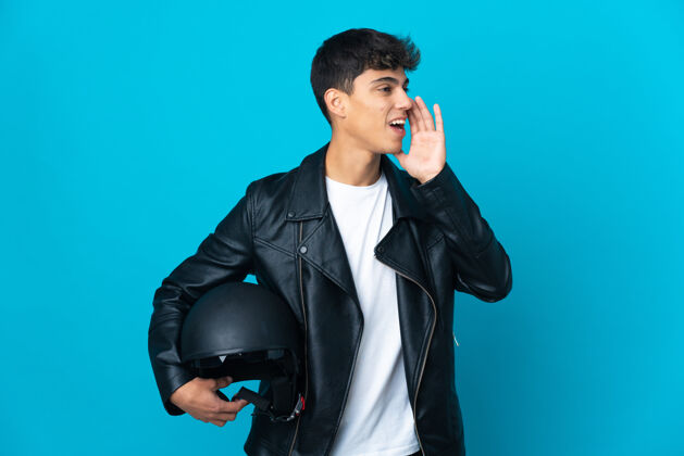 情绪一个戴着摩托车头盔的年轻人站在隔离的蓝色墙壁上 张大嘴巴向一边喊叫声音信息夹克