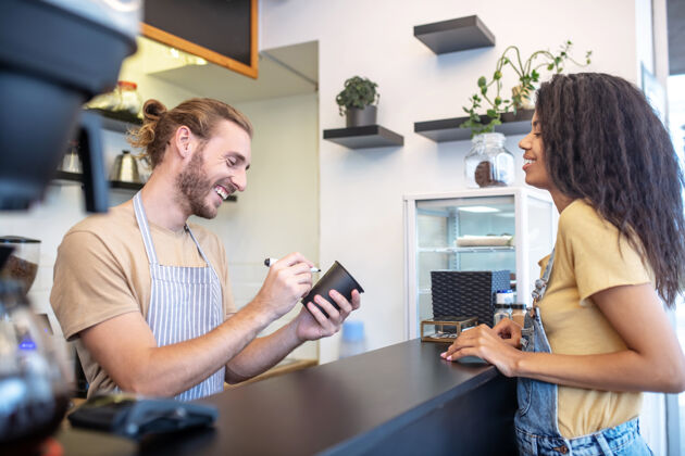 创业客户 咖啡馆大笑年轻的留着胡子的男人拿着马克笔和玻璃杯 长头发的女人在咖啡馆的柜台旁点菜咖啡家具咖啡机