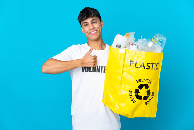微笑拿着装满塑料瓶的袋子翻过蓝色的墙壁 竖起大拇指志愿者男性积极