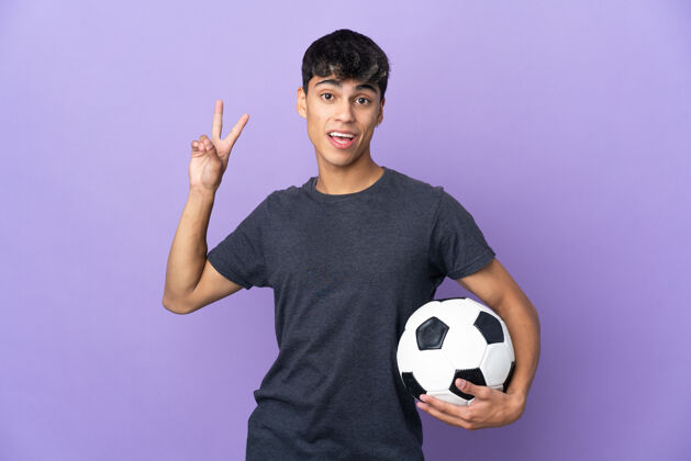 运动装年轻的足球运动员男子在孤立的紫色背景微笑着 并显示胜利的标志快乐运动员是的