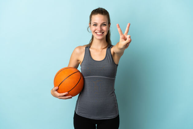 年轻在蓝色背景上打篮球的年轻女子面带微笑 露出胜利的标志健身身体是