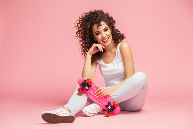 时髦快乐迷人的年轻女子坐在粉红色的滑板上欢乐滑板衣服