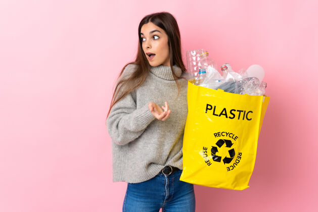 环境年轻的白种女人拿着一个装满塑料瓶的袋子在粉红色的背景下 一边看着一边惊讶的表情责任年轻惊喜