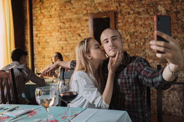 手机恋爱中的男女在餐厅合影——一位迷人的白种人一边拿着智能手机自拍 一边亲吻男友的脸颊手男人欢呼