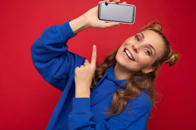 哇迷人的微笑的年轻金发女子穿着时尚的蓝色连帽衫隔离在红色背景上手机学生通讯