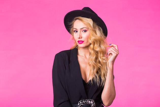 样式年轻漂亮的金发女郎戴着黑色时髦的帽子在粉红色的墙上摆姿势皮肤嘴唇护理
