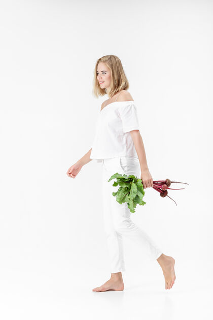 人一位穿着白色上衣的金发美女手持一根长着绿叶的甜菜根背景.健康还有维生素年轻背景夏天
