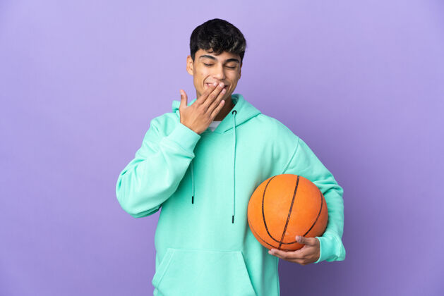 比赛在孤立的紫色背景上打篮球的年轻人开心地微笑着用手捂住嘴比赛大笑微笑