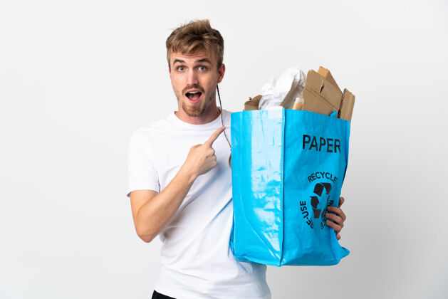 废物年轻的金发男子拿着一个回收袋 里面装满了回收纸 孤立地站在白色的一边惊讶地指着垃圾生态责任