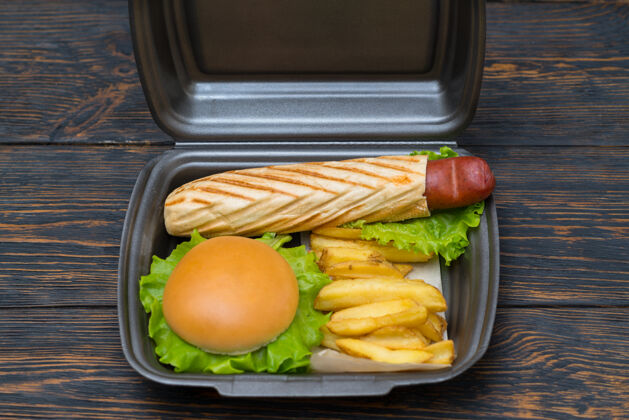 沙拉外卖香肠狗在一个烤制的法式面包 汉堡和薯条在一个一次性聚苯乙烯容器高角度上菜肉饼
