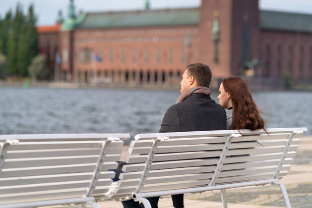 秋天一对年轻的夫妇在长凳上放松 享受日落 因为他们可以俯瞰水和历史建筑在一个城市复制空间寒冷历史放松
