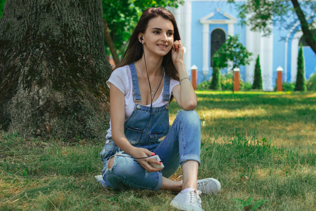 电话快乐的年轻黑发少年坐在草地上 用耳机听音乐公园成人情绪